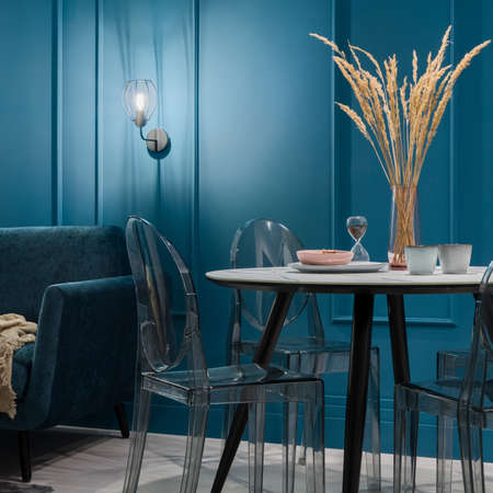 Modern dining table with stylish plastic chairs in nice living room with teal blue walls with decorative molding and lightingの写真素材