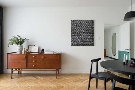Stylish and vintage, wooden sideboard with decorations in bright room with modern art, black, round dining table with chairs and wooden floorの写真素材