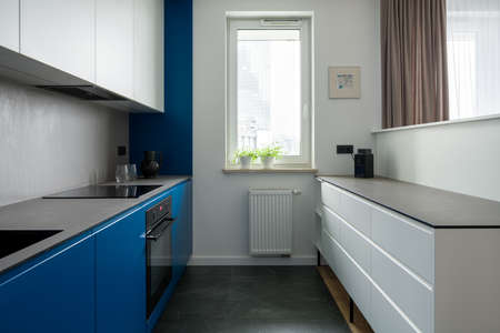 Narrow and modern kitchen with window, white drawers, gray countertops and stylish blue furniture and wallの写真素材