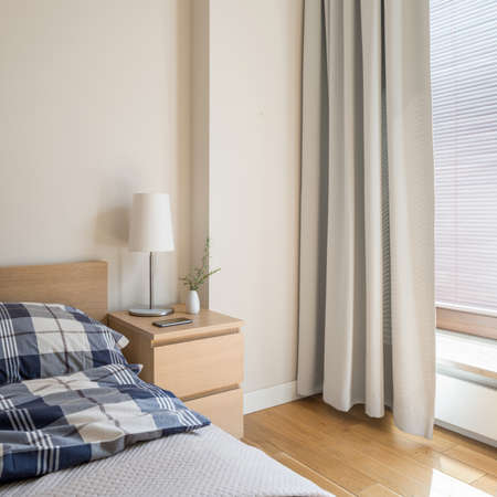 Simple wooden bedside table with white lamp and decorations next to bed in bright bedroomの写真素材