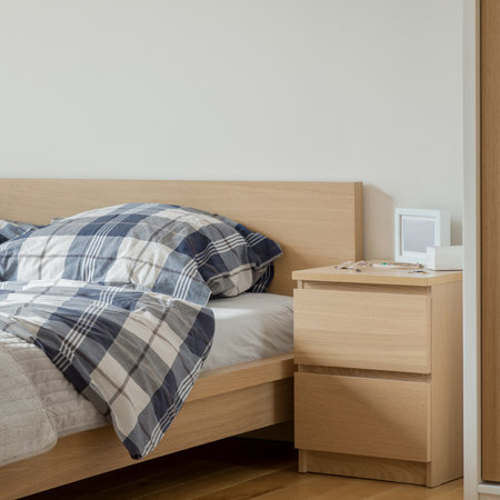 Wooden bedside table with two drawers next to comfortable bed with wooden frame and headboard in simple bedroomの写真素材