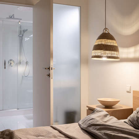 Cozy master bedroom with rattan lamp on and open doors to bathroom with showerの写真素材