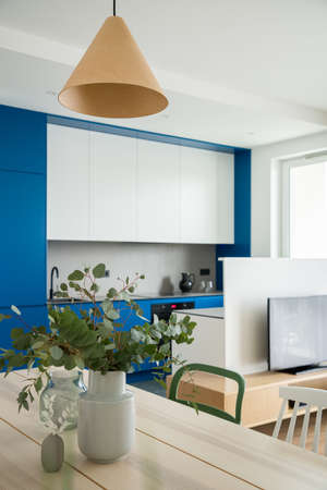 Stylish wooden dining table under decorative lamp next to modern kitchen with white and blue cupboards open to tv roomの写真素材