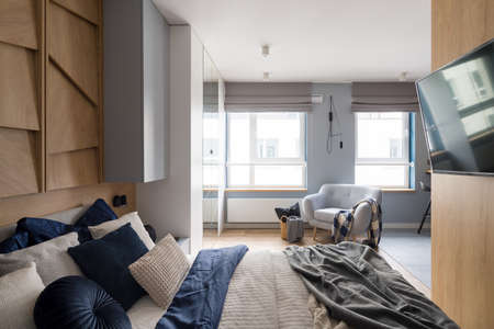 Stylish bedroom with cozy bed and nice bedclothes, big windows, comfortable armchair and tv on wooden wallの写真素材