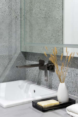 Small, white bathroom washbasin with black tap and decorative vase with dried grass and soap bar on gray countertop and gray tilesの写真素材
