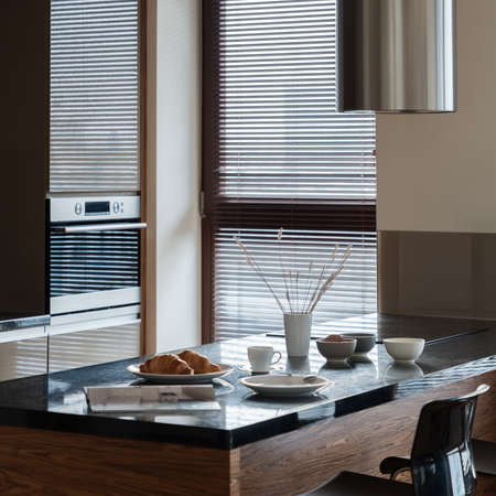 Close-up on breakfast on kitchen island with black countertop with induction hob under silver kitchen hood and with black chair in small kitchen with windowの写真素材