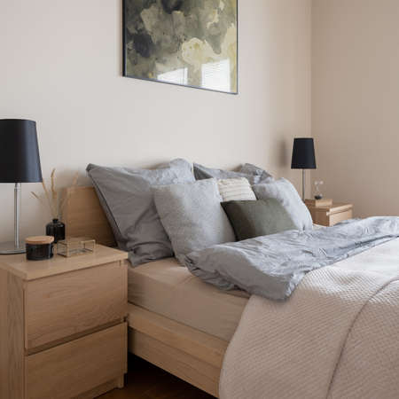 Close-up on comfortable bed, two wooden nightstands with black lamps and decorations in bright bedroomの写真素材