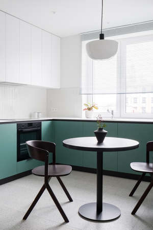 Modern dining table with two chairs in stylish kitchen with big window, white and green cupboards and terrazzo floorの写真素材