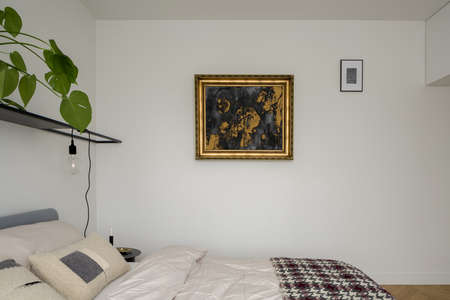 Nice painting in gold frame on white wall in bedroom with simple bed and black shelf with plant and lampの写真素材