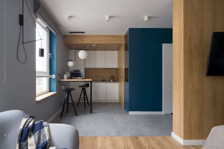 Small and modern kitchen with white cupboards, wooden wall and table open to room with cozy armchairの写真素材