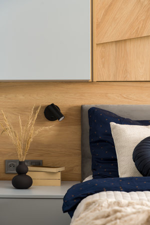 Black vase with dried grass and books on gray bedside table next to comfortable bed with stylish bedclothesの写真素材