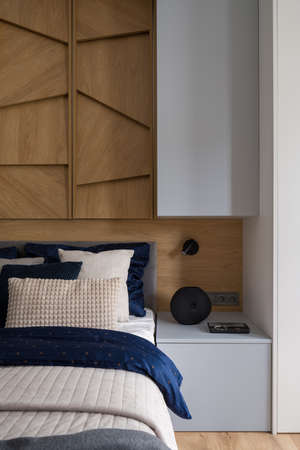 Gray bedside table next to stylish decorated bed in bright bedroom with wooden decorations on wallの写真素材