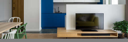 Panorama of simple tv room with wooden sideboard open to dining area with table and eclectic white and green chairs open to modern kitchen with white and blue furnitureの写真素材