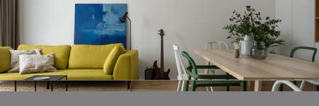 Panorama of small and stylish living room with comfortable, modern yellow sofa and nice black coffee table and dining area with wooden table and different chairsの写真素材