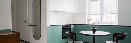 Panorama of stylish apartment with modern white and mint green kitchen cupboards with black dining table and chairs open to corridor with trendy oval mirrorの写真素材