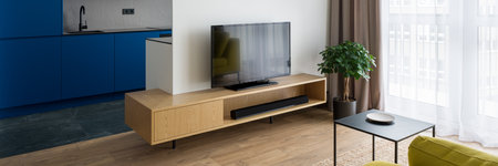 Panorama of bright room with tv on wooden sideboard and big window next to modern kitchen with navy blue furnitureの写真素材