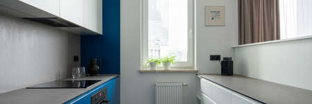 Panorama of narrow and modern kitchen with window, gray countertops and stylish blue and white furniture and wallの写真素材