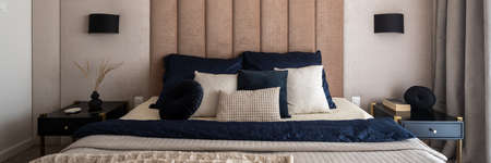 Panorama of elegant bedroom with upholstered, beige wall behind comfortable and decorated bed and with two, black bedside tables with decorationsの写真素材
