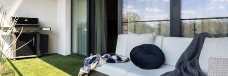 Panorama of spacious terrace with gas grill, comfortable, rattan sofa and floor with fake grass and balcony window openの写真素材