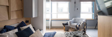 Panorama of bright bedroom with cozy bed and nice bedclothes, big windows, tv on wooden wall and comfortable armchairの写真素材