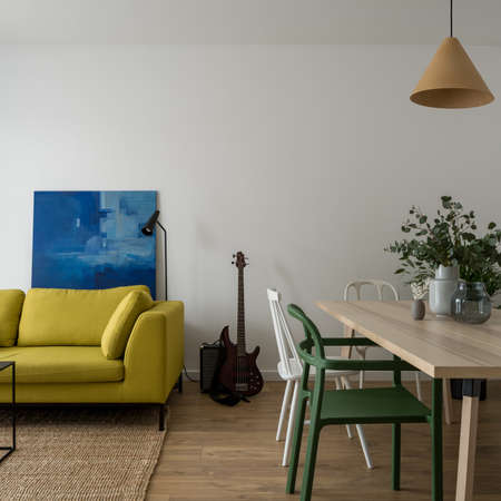 Simple living room with comfortable, yellow sofa, modern art and guitar and dining area with wooden table and white and green chairsの写真素材