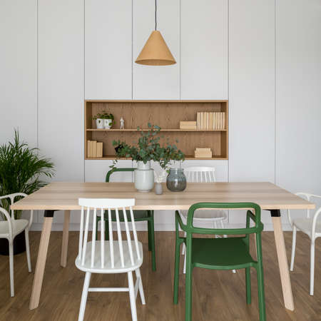 Stylish dining room with simple, wooden table and various white and green chairs, small lamp and modern wardrobe wall with wooden shelfの写真素材