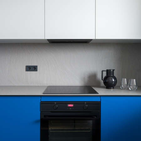 Close-up on simple black oven and induction hob in minimalist kitchen with white and blue cupboards and gray countertop with backsplashの写真素材