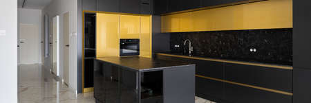 Panorama of spacious and luxury kitchen with black and golden cupboards and stylish kitchen island with black countertop next to white corridor with marble floorの写真素材