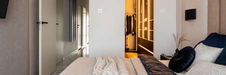 Panorama of elegant designed bedroom with big walk in closet with led light, comfortable bed and open doors to corridorの写真素材