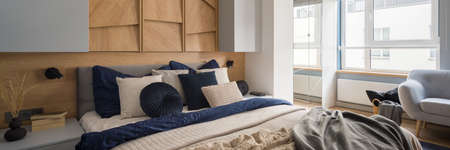Panorama of spacious and cozy bedroom with decorative, wooden wall, gray nightstands and cupboards, big window and comfortable bed with stylish pillows and bedclothesの写真素材