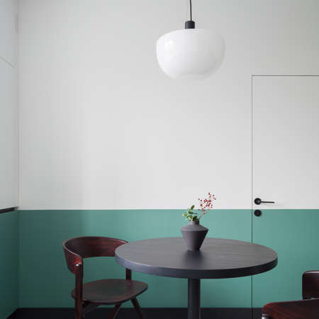 Stylish, round, wooden and dark dining table with decorative vase and modern chairs under new lamp in room with white and green wallの写真素材