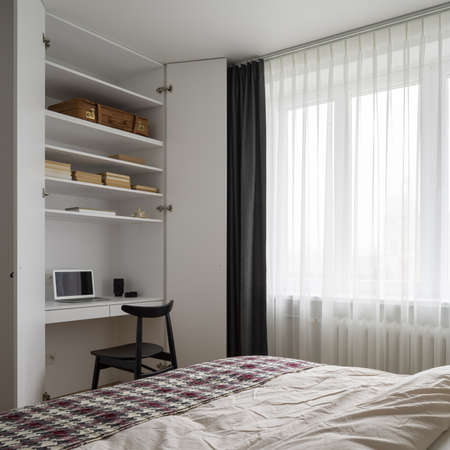 Simple bedroom with comfortable bed and big window behind curtains and stylish desk to work in white wardrobeの写真素材