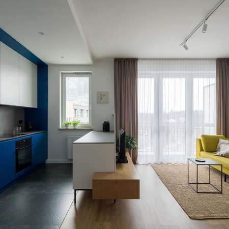 Contemporary open plan apartment interior with modern designed kitchen with white and blue cupboards open to stylish living room with big windowsの写真素材