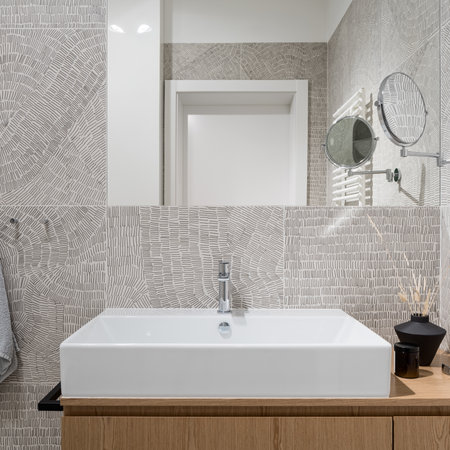 Close-up on stylish, white washbasin with wooden cabinet in new bathroom with modern, patterned tiles and big mirrorの写真素材