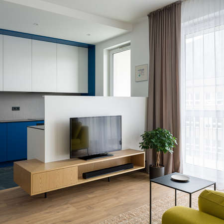Small living room with tv on wooden sideboard and big window open to modern kitchen with white and blue furnitureの写真素材