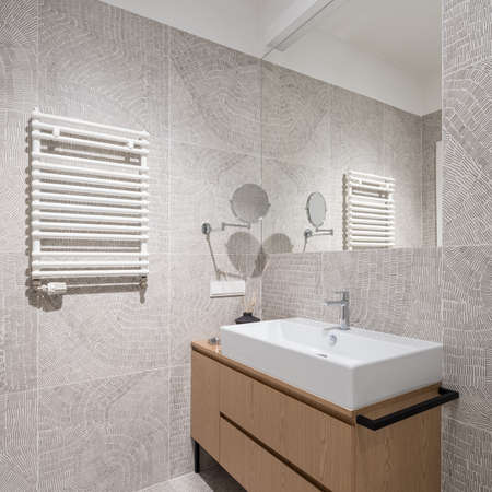 Small bathroom with big mirror above white washbasin with wooden cabinet and with modern and trendy wall tiles with stylish patternの写真素材