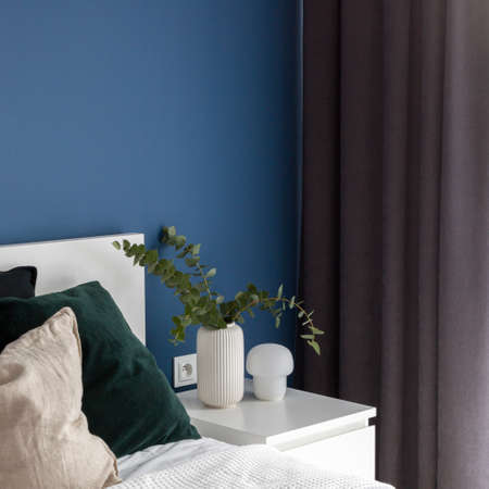 Close-up on simple white bedside table with decorative lamp and vase with eucalyptus next to cozy bed in bedroom with blue wallの写真素材