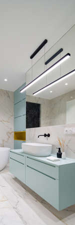 Vertical panorama of elegant bathroom with marble floor and wall tiles with modern blue cabinets and drawers under washbasin and led ceiling lightsの写真素材