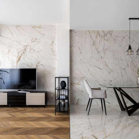 Stylish dining area with modern table and comfortable chair and marble wall and floor tiles next to tv room with big screen and wooden floorの写真素材