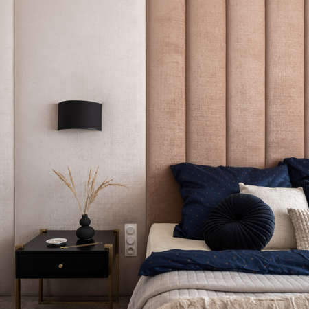 Close-up on stylish, black bedside table with decorations under black lamp next to comfortable bed with fancy bedclothes and with upholstered wallの写真素材