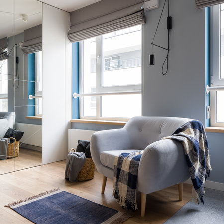 Cozy and gray armchair with blanket in bright room with big windows and wardrobe with mirror doorの写真素材