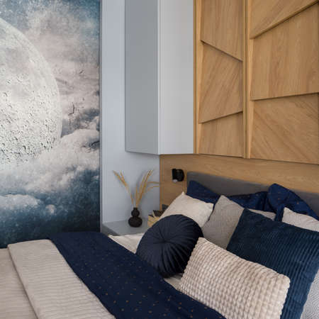 Decorative bedclothes on cozy bed in small bedroom with wooden decor on the wall and wallpaper with moonの写真素材