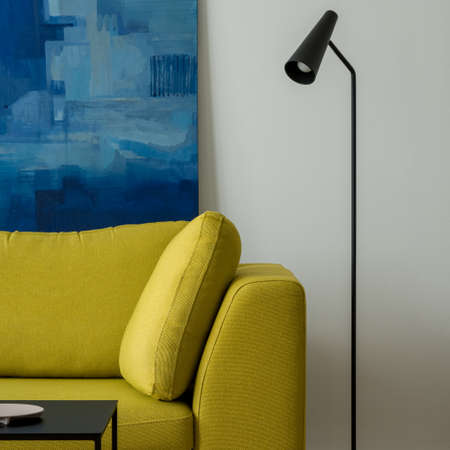 Close-up on modern, yellow couch next to simple, black lamp in room with white walls and artの写真素材