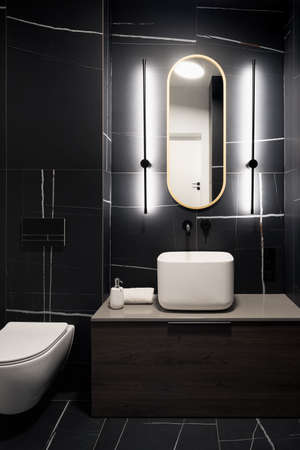 Stylish dark bathroom with black tiles, modern square washbasin and oval mirror with led lightの写真素材