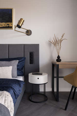 White bedside table in bedroom with cozy bed and wooden desk with chairの写真素材