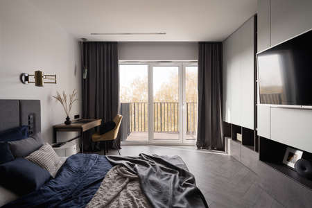 Spacious bedroom with comfortable bed, big tv, desk with stylish chair and window with doors to balconyの写真素材
