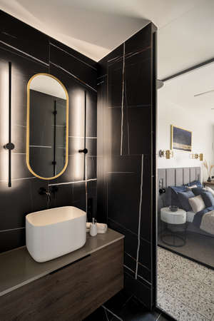 Led lights by oval mirror over square washbasin in modern bathroom with black, marble tiles and glass wall to bedroomの写真素材