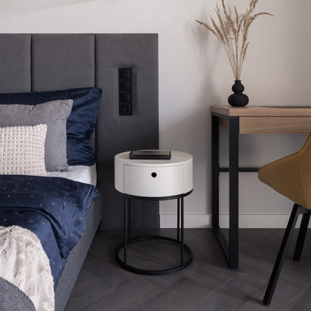 Close-up of stylish and white bedside table in bedroom with cozy bed and wooden desk with chair