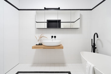 Spacious bathroom in white and black tiles, freestanding bathtub with black tap and mirrored cabinet over washbasin in wooden shelfの写真素材