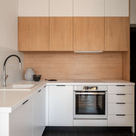 Small and simple kitchen with white and wooden cupboards, countertops, drawers, oven and sink with black tapの写真素材